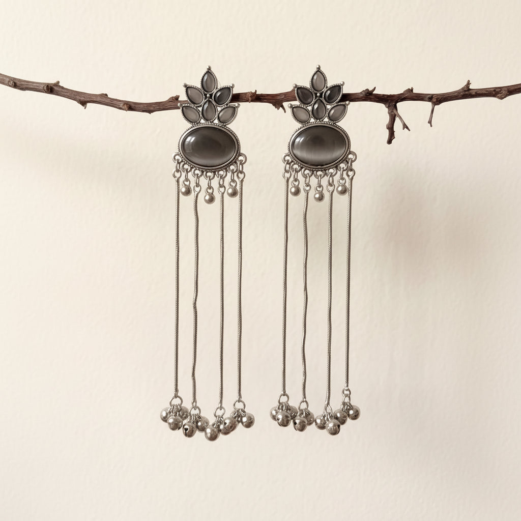 A pair of silver-toned dangle earrings with dark oval stones and dangling chain bells, hanging from a small branch against a plain light beige wall.