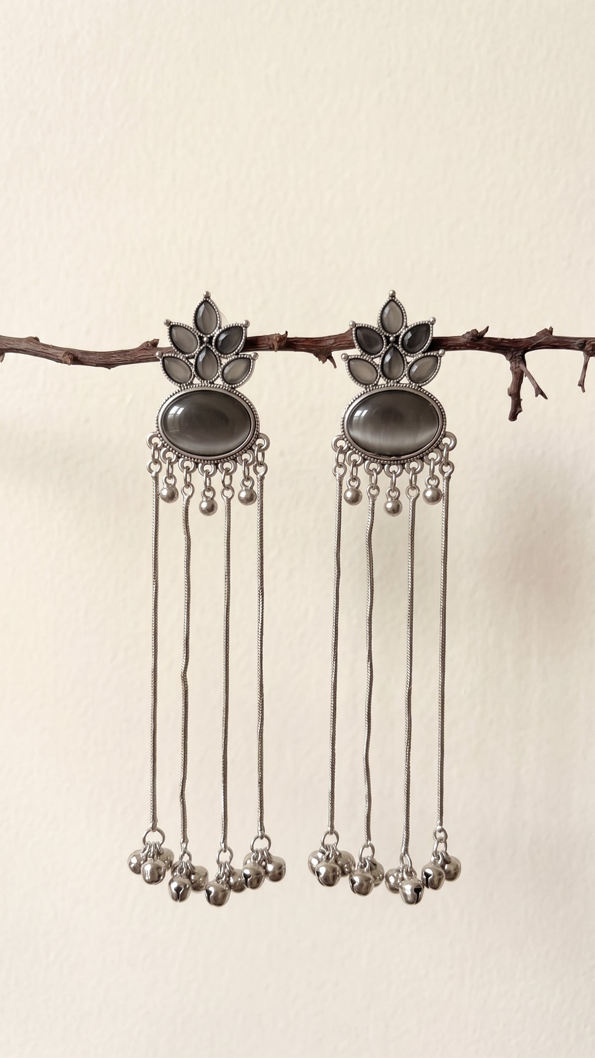 A pair of silver-toned dangle earrings with dark oval stones and dangling chain bells, hanging from a small branch against a plain light beige wall.