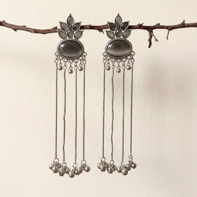 A pair of silver-toned dangle earrings with dark oval stones and dangling chain bells, hanging from a small branch against a plain light beige wall.