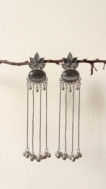 A pair of silver-toned dangle earrings with dark oval stones and dangling chain bells, hanging from a small branch against a plain light beige wall.