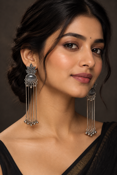 A close-up portrait of a woman wearing ornate silver-toned dangle earrings with oval gray centers and dangling bead chains, against a dark brown studio background.