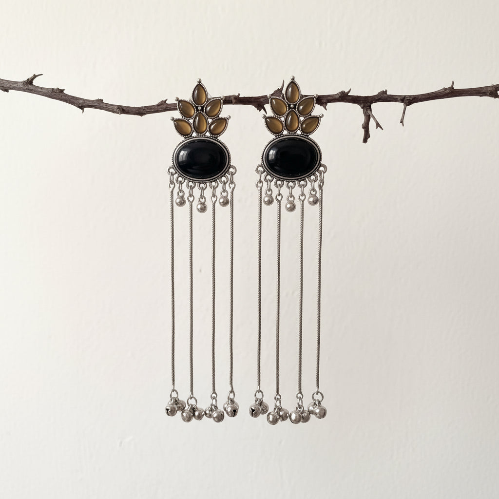 A pair of silver-toned dangle earrings with oval black stones and gold-toned floral details, hanging from a thin branch against a plain off-white wall.