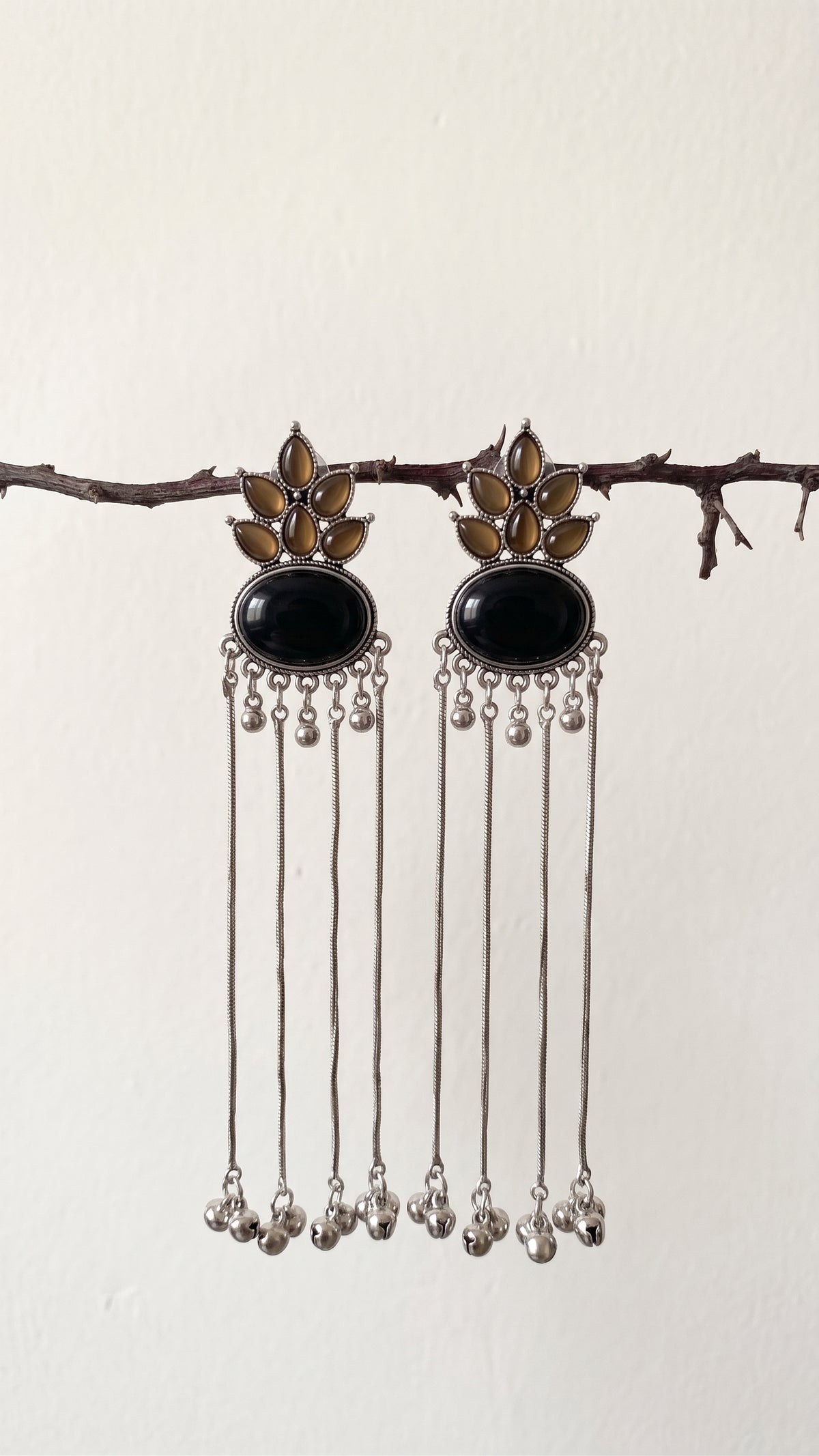 A pair of silver-toned dangle earrings with oval black stones and gold-toned floral details, hanging from a thin branch against a plain off-white wall.