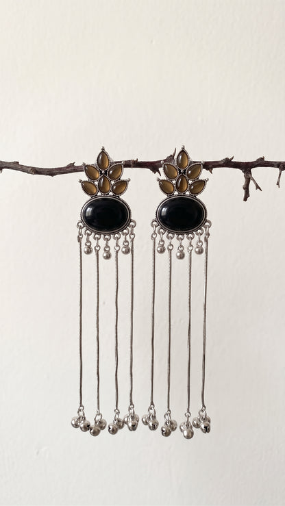 A pair of silver-toned dangle earrings with oval black stones and gold-toned floral details, hanging from a thin branch against a plain off-white wall.