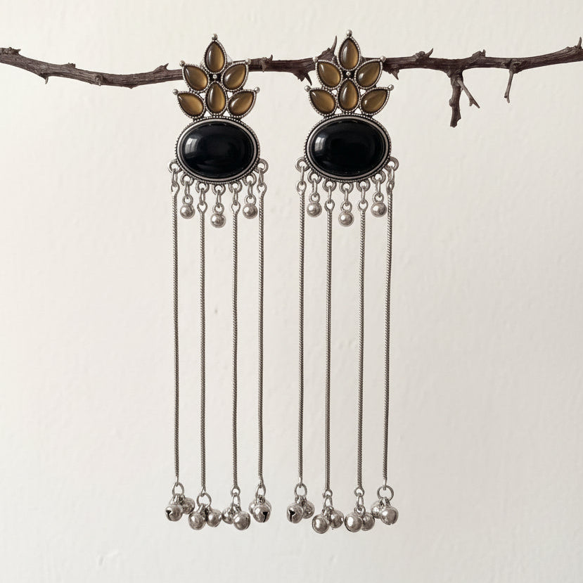 A pair of silver-toned dangle earrings with oval black stones and gold-toned floral details, hanging from a thin branch against a plain off-white wall.