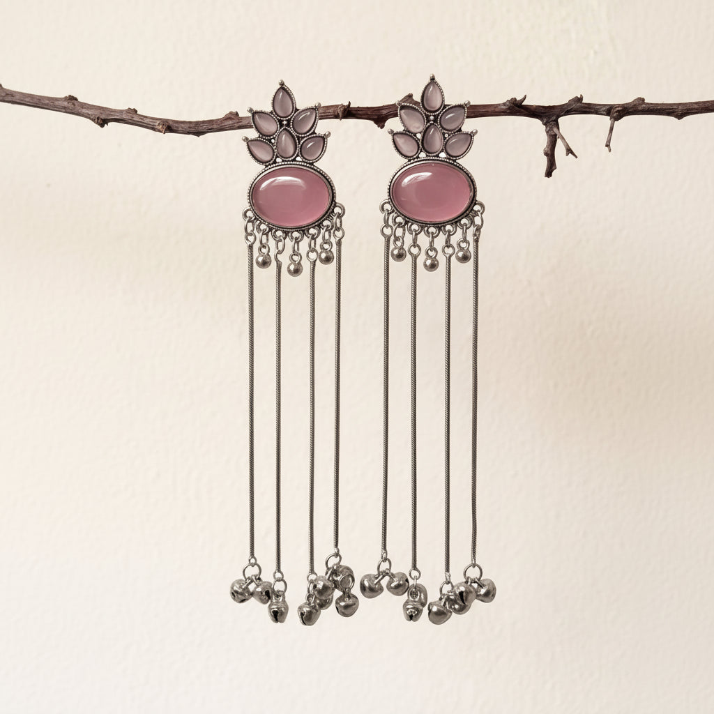 A pair of silver-toned dangle earrings with oval pink stone centers and dangling silver bells, hanging from a thin branch against a plain off-white wall.