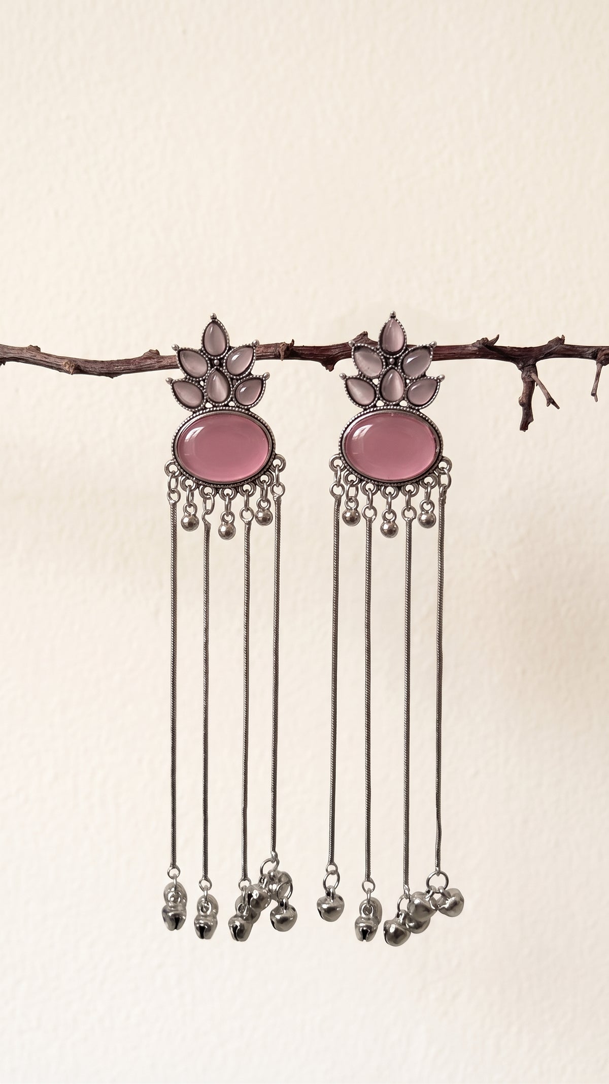 A pair of silver-toned dangle earrings with oval pink stone centers and dangling silver bells, hanging from a thin branch against a plain off-white wall.