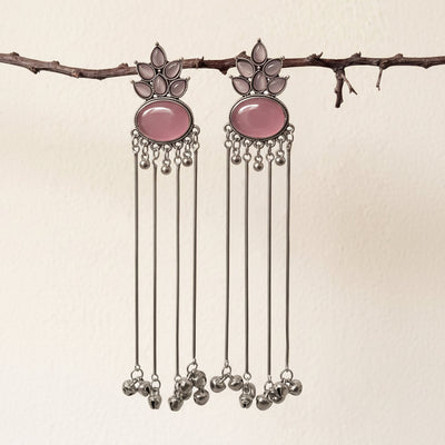 A pair of silver-toned dangle earrings with oval pink stone centers and dangling silver bells, hanging from a thin branch against a plain off-white wall.
