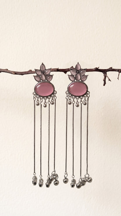 A pair of silver-toned dangle earrings with oval pink stone centers and dangling silver bells, hanging from a thin branch against a plain off-white wall.