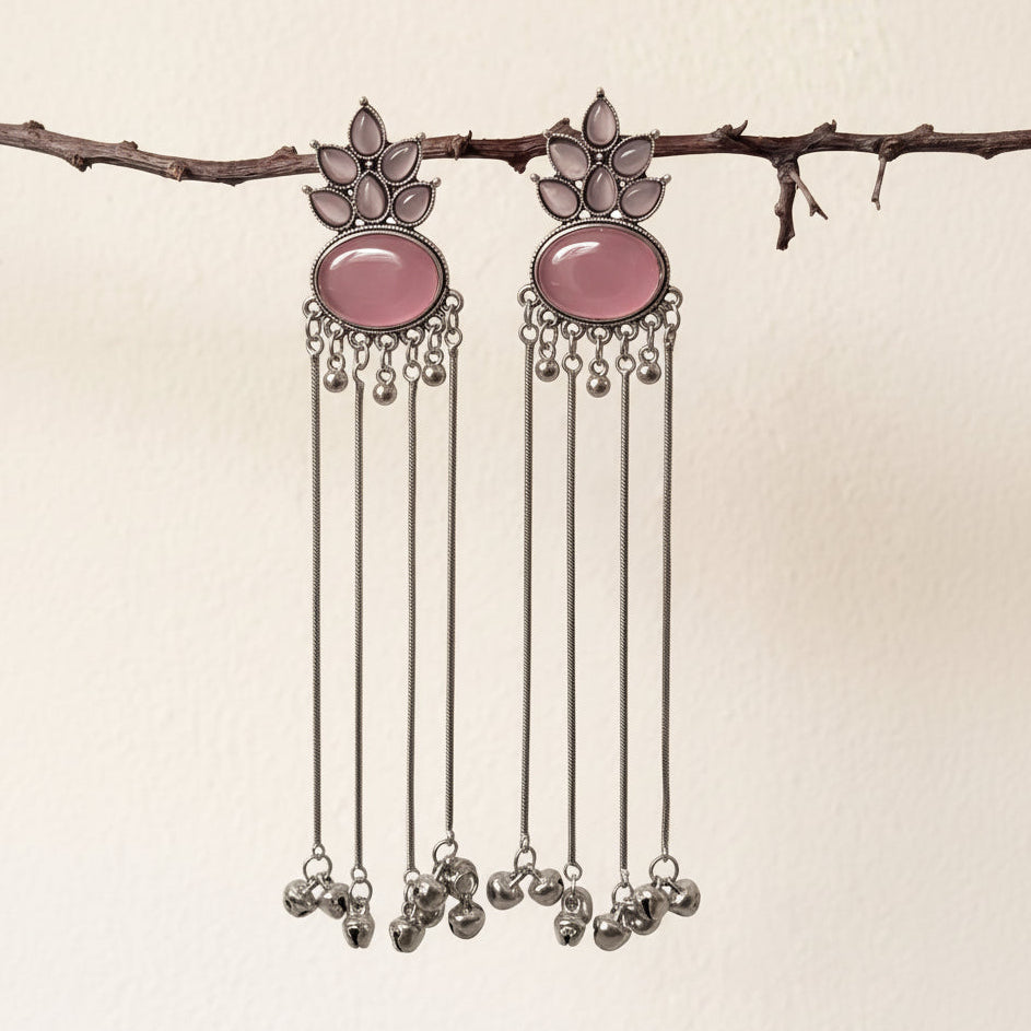 A pair of silver-toned dangle earrings with oval pink stone centers and dangling silver bells, hanging from a thin branch against a plain off-white wall.