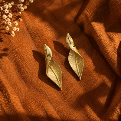 A pair of gold-toned leaf-shaped earrings displayed on draped rust-orange fabric with dried flowers in the corner.