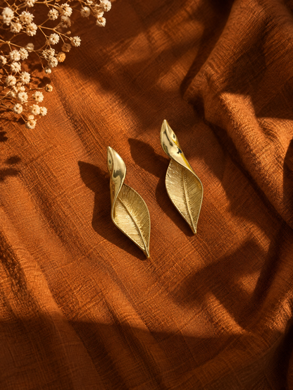 A pair of gold-toned leaf-shaped earrings displayed on draped rust-orange fabric with dried flowers in the corner.