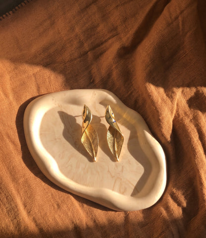 A pair of gold-toned leaf-shaped drop earrings resting on an organic-shaped cream ceramic tray, set against a draped brown fabric background with warm sunlight and shadows.