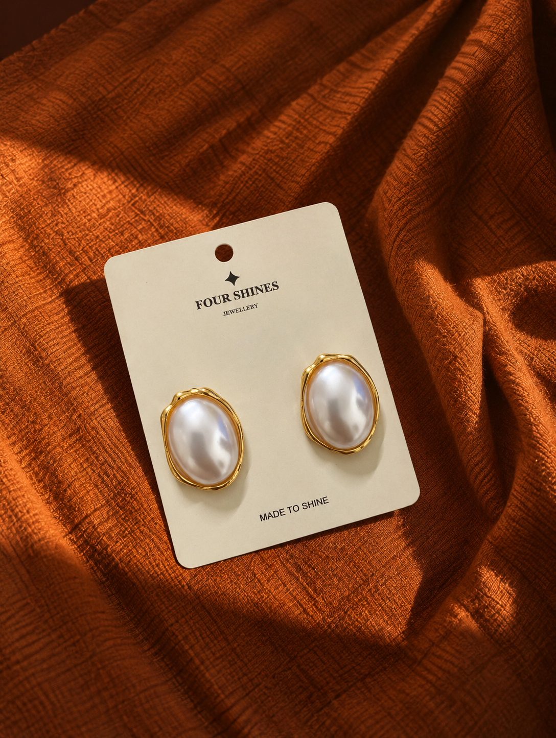 A pair of oval pearl stud earrings with gold bezel settings, mounted on a cream-colored card labeled 'FOUR SHINES JEWELRY', placed on a draped rust-orange fabric background.