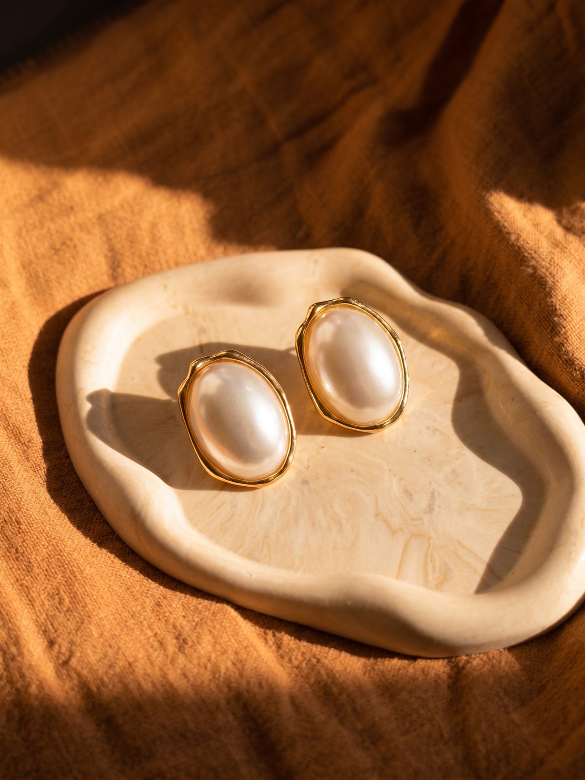 A pair of gold-tone stud earrings with large oval white pearl-like centers, displayed on a cream ceramic dish over a warm brown fabric background under dramatic sunlight.