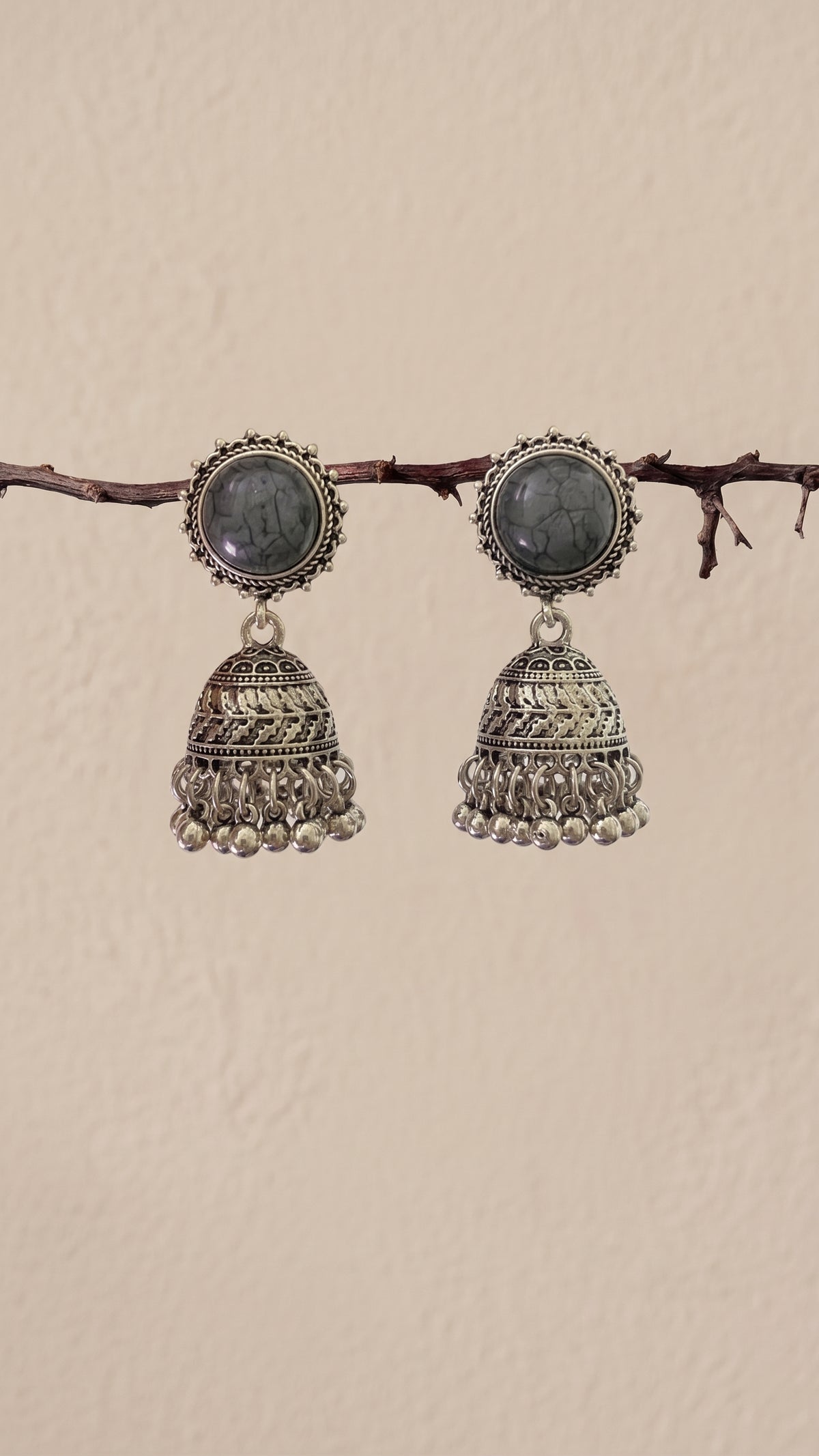 A pair of traditional Indian silver jhumka earrings with oval dark gray stones, hanging from a thin brown branch against a textured beige wall.