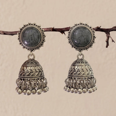 A pair of traditional Indian silver jhumka earrings with oval dark gray stones, hanging from a thin brown branch against a textured beige wall.