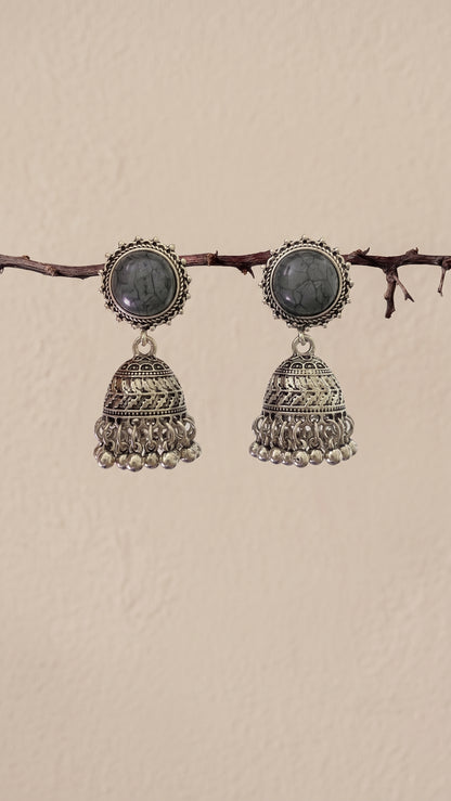 A pair of traditional Indian silver jhumka earrings with oval dark gray stones, hanging from a thin brown branch against a textured beige wall.