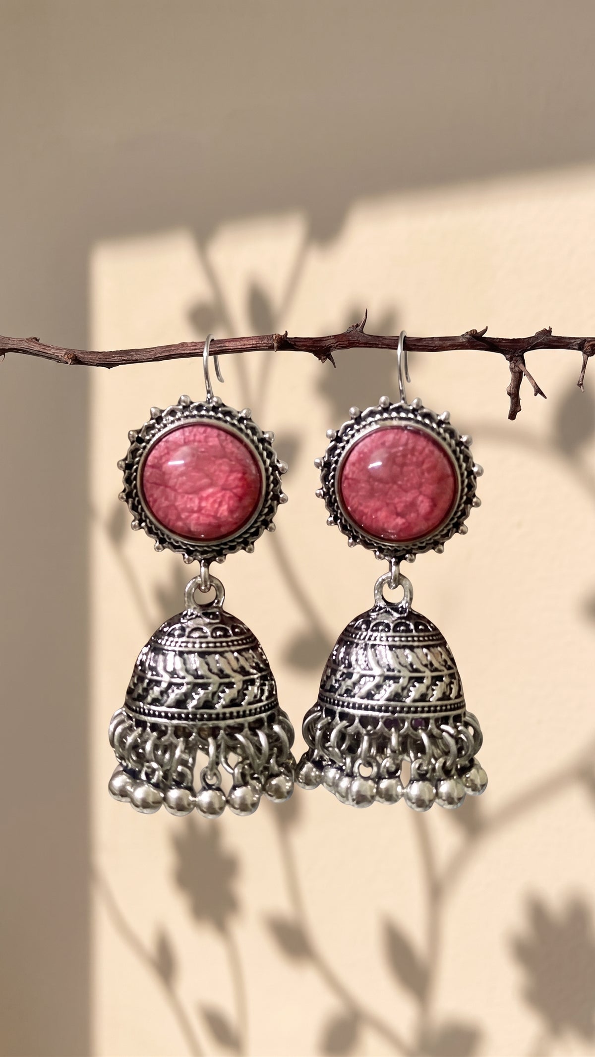 A pair of silver-toned Indian jhumka earrings with round pink cabochon stones, hanging from a thin brown branch against a beige wall with leaf shadows.