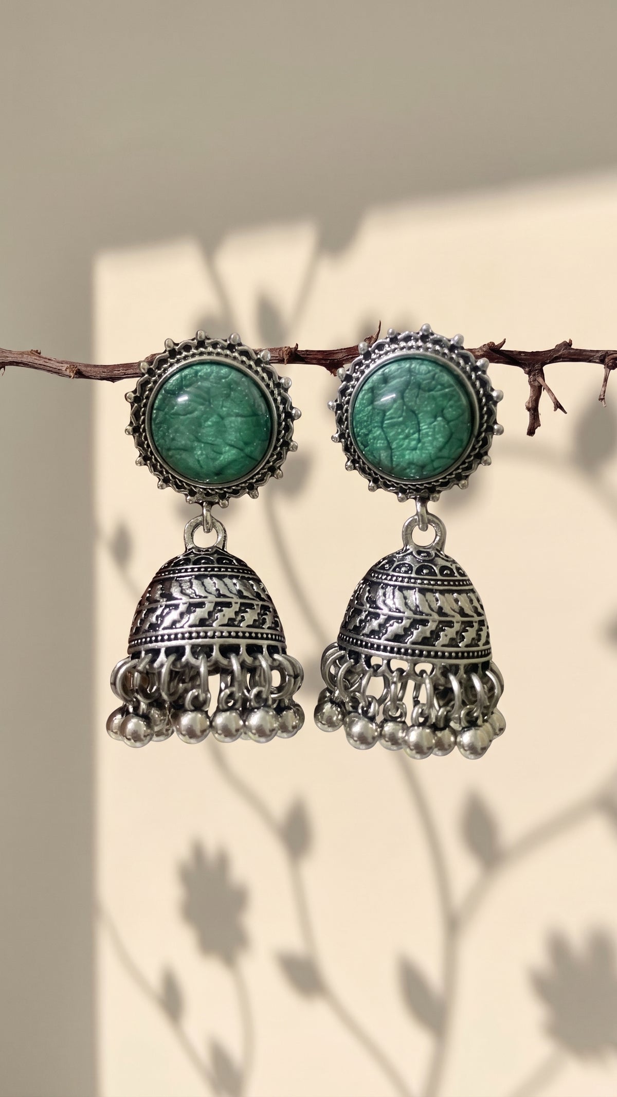 A pair of traditional Indian silver jhumka earrings with green stone studs, hanging from a thin branch against a warm beige background with soft leaf shadows.