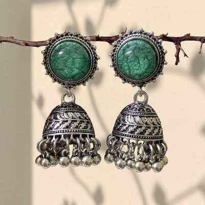 A pair of traditional Indian silver jhumka earrings with green stone studs, hanging from a thin branch against a warm beige background with soft leaf shadows.
