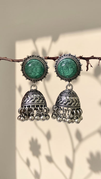 A pair of traditional Indian silver jhumka earrings with green stone studs, hanging from a thin branch against a warm beige background with soft leaf shadows.