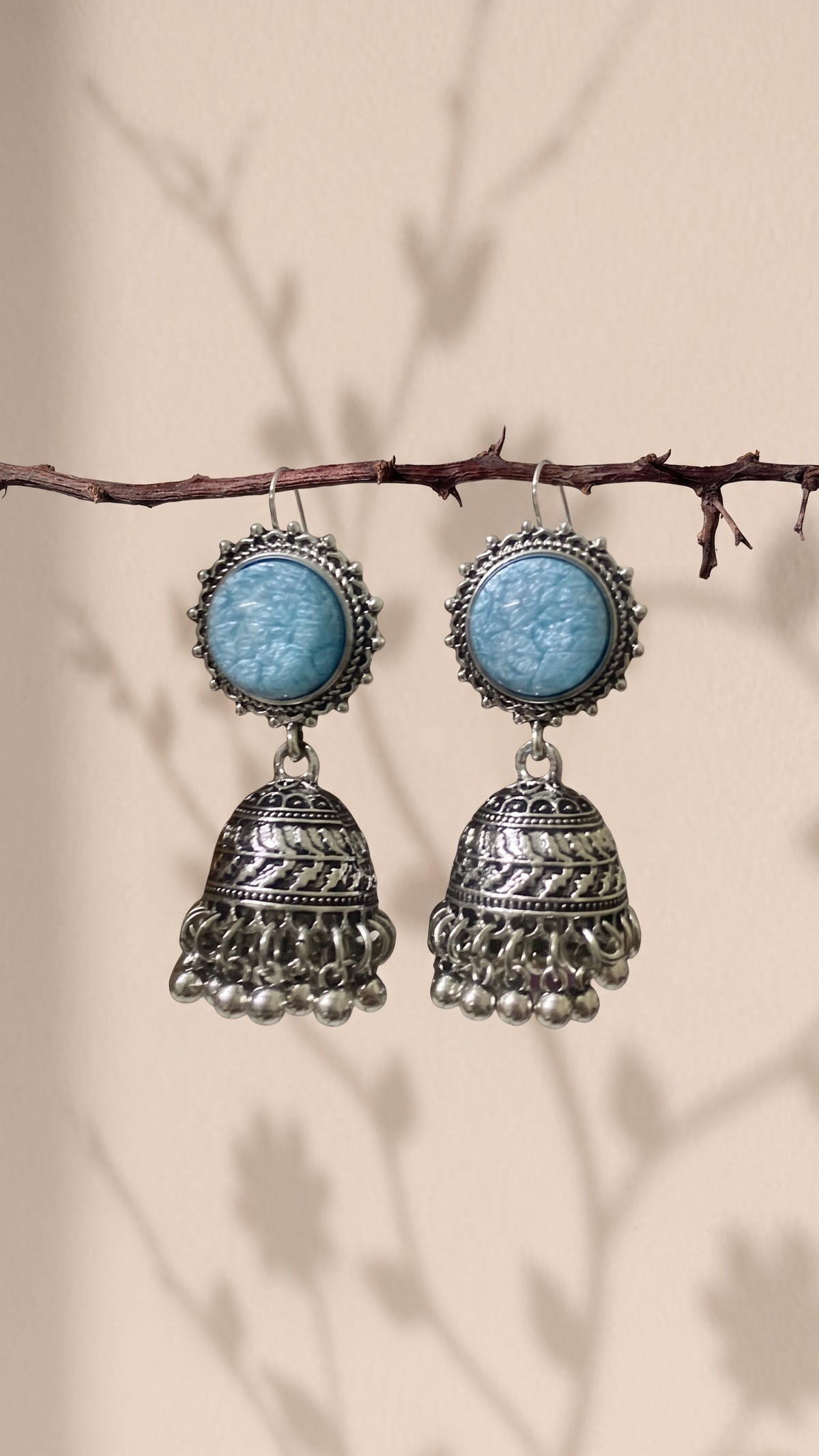 A pair of bohemian silver jhumka earrings with light blue cabochon stones, hanging from a thin brown branch against a beige background with soft leaf shadows.