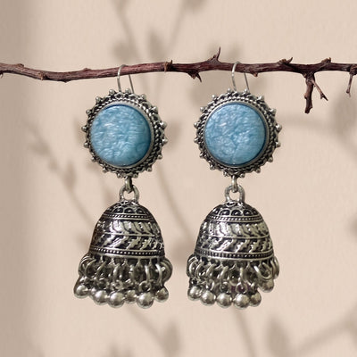 A pair of bohemian silver jhumka earrings with light blue cabochon stones, hanging from a thin brown branch against a beige background with soft leaf shadows.