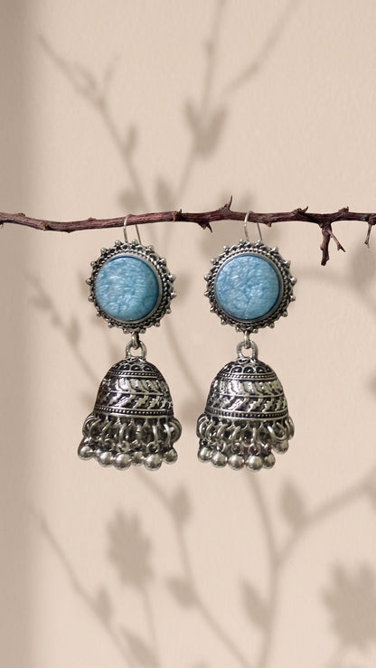A pair of bohemian silver jhumka earrings with light blue cabochon stones, hanging from a thin brown branch against a beige background with soft leaf shadows.
