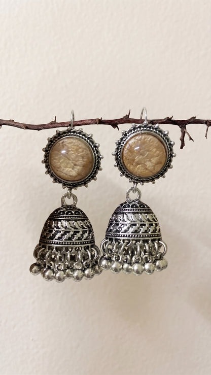 A pair of traditional Indian silver-toned jhumka earrings with oval beige stone inlays, hanging from a small branch against a plain beige wall.