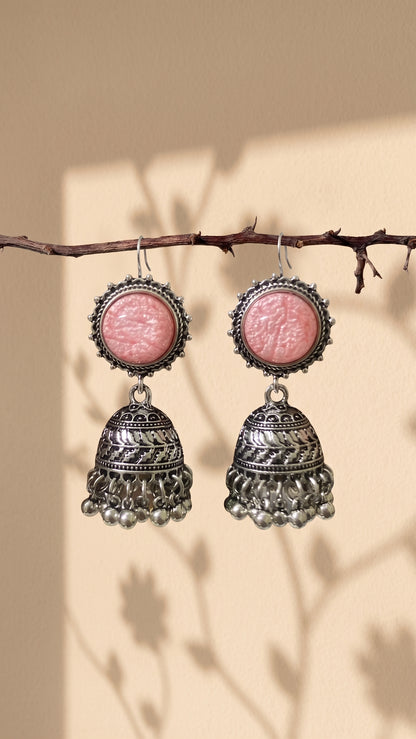 A pair of silver-toned jhumka earrings with round pink cabochon stones, hanging from a thin branch against a warm beige background with soft leaf shadows.