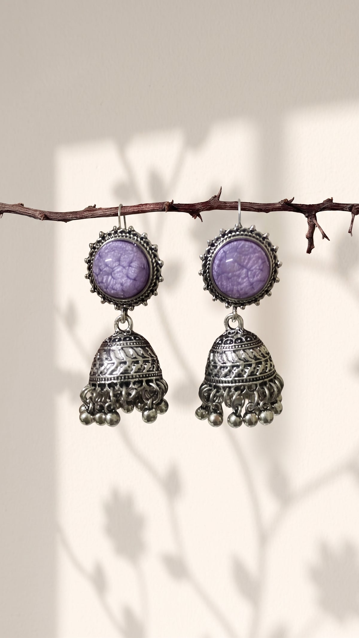 A pair of silver-toned jhumka earrings with oval purple stone tops, hanging from a thin branch against a warm beige background with soft leaf shadows.