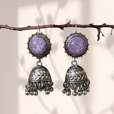 A pair of silver-toned jhumka earrings with oval purple stone tops, hanging from a thin branch against a warm beige background with soft leaf shadows.